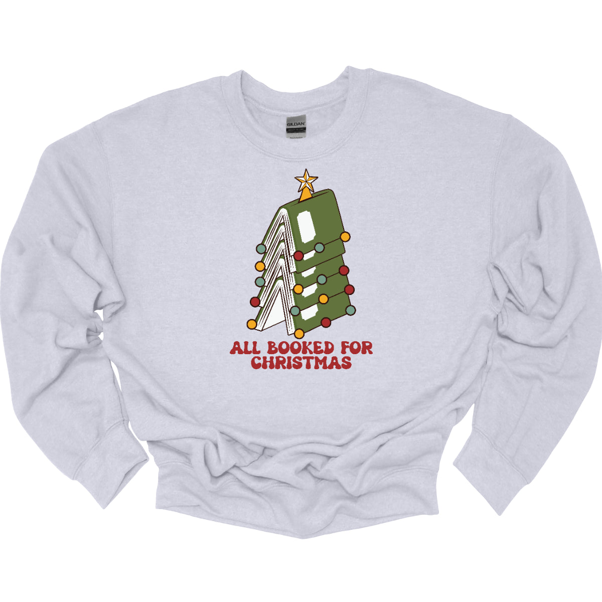 This whimsical t-shirt features the playful phrase "All Booked for Christmas" in a bold, red bubbly font that captures the festive spirit. Above the text, a delightful image showcases a stack of green books, artfully arranged to resemble a decorated Christmas tree. Shop Gorgeousware.com
