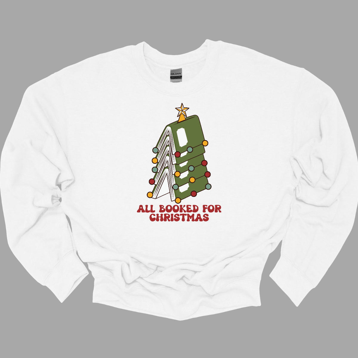 This whimsical t-shirt features the playful phrase "All Booked for Christmas" in a bold, red bubbly font that captures the festive spirit. Above the text, a delightful image showcases a stack of green books, artfully arranged to resemble a decorated Christmas tree. Shop Gorgeousware.com