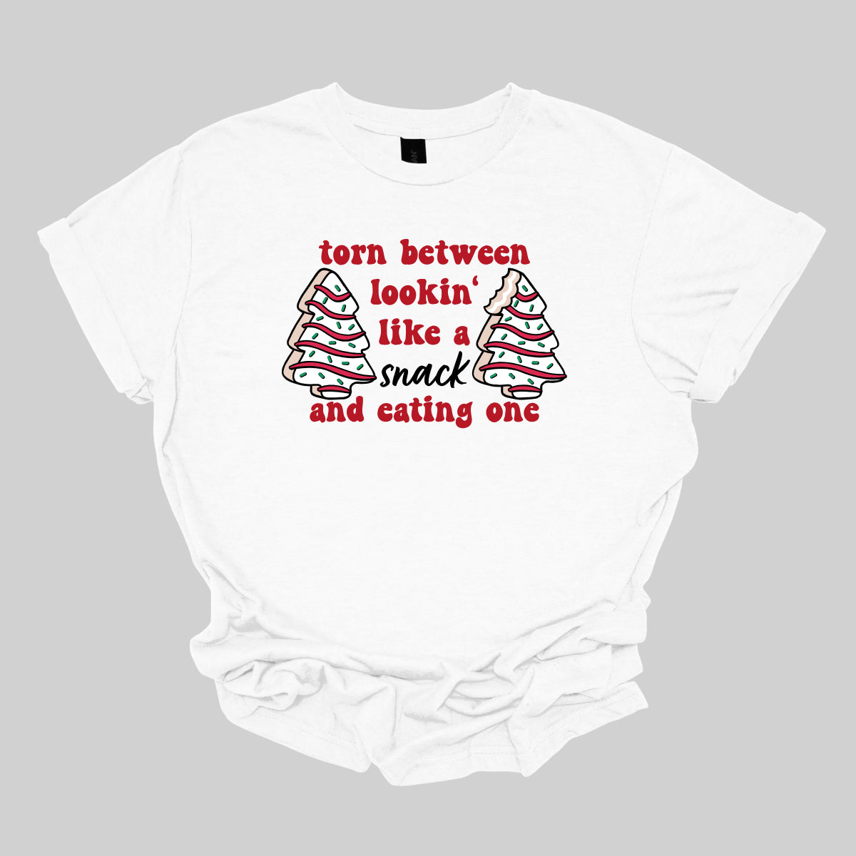 This fun t-shirt features the playful phrase "Torn between lookin' like a snack and eating one" in a vibrant red bubbly font, adding a lively and festive touch. The word "snack" stands out in an elegant black cursive font, creating a nice contrast. Flanking the text are adorable illustrations of Christmas tree cakes on each side, enhancing the holiday spirit and adding a whimsical flair. This shirt is perfect for anyone who loves to celebrate the season with humor and style! Shop Gorgeousware.com