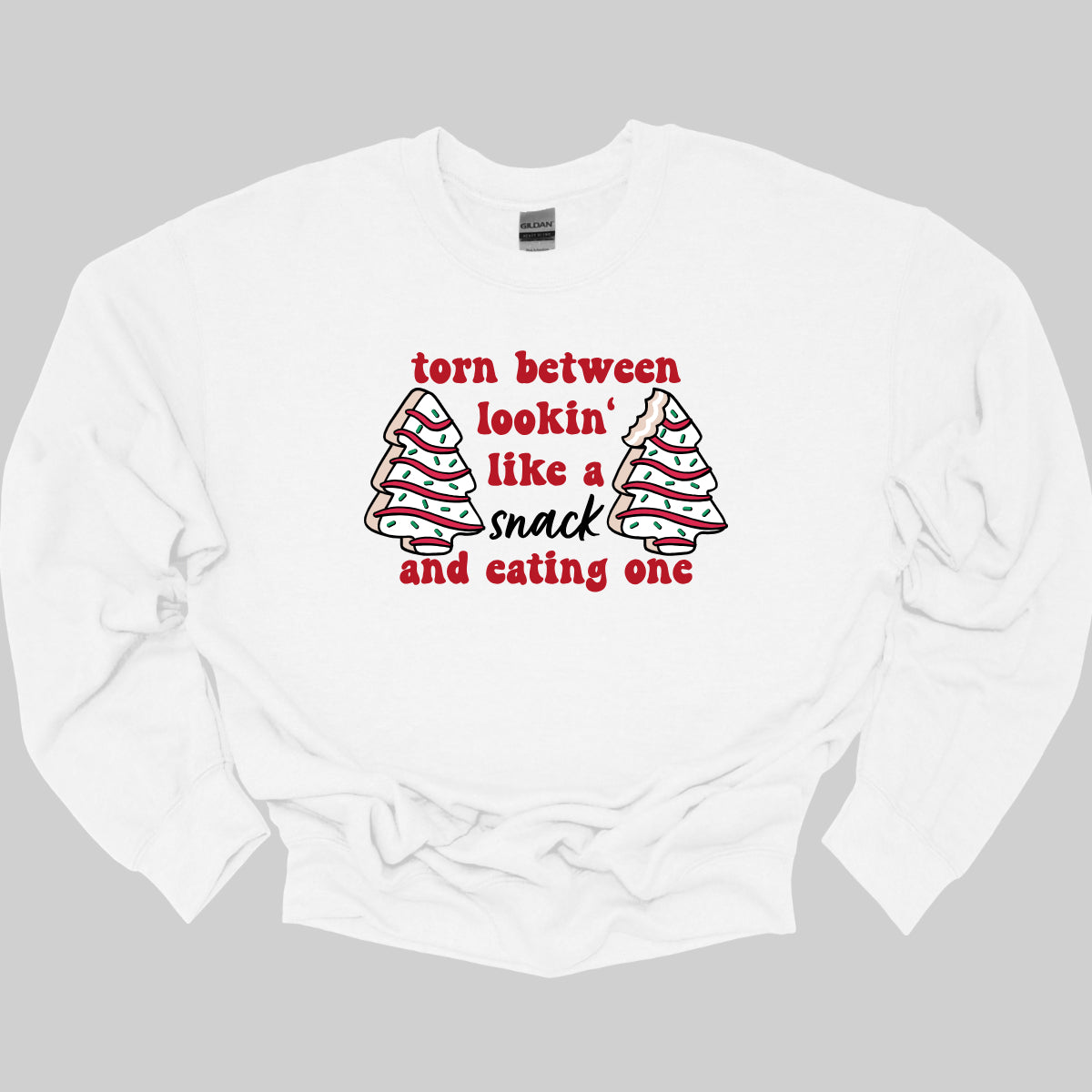 This fun t-shirt features the playful phrase "Torn between lookin' like a snack and eating one" in a vibrant red bubbly font, adding a lively and festive touch. The word "snack" stands out in an elegant black cursive font, creating a nice contrast. Flanking the text are adorable illustrations of Christmas tree cakes on each side, enhancing the holiday spirit and adding a whimsical flair. This shirt is perfect for anyone who loves to celebrate the season with humor and style! Shop Gorgeousware.com