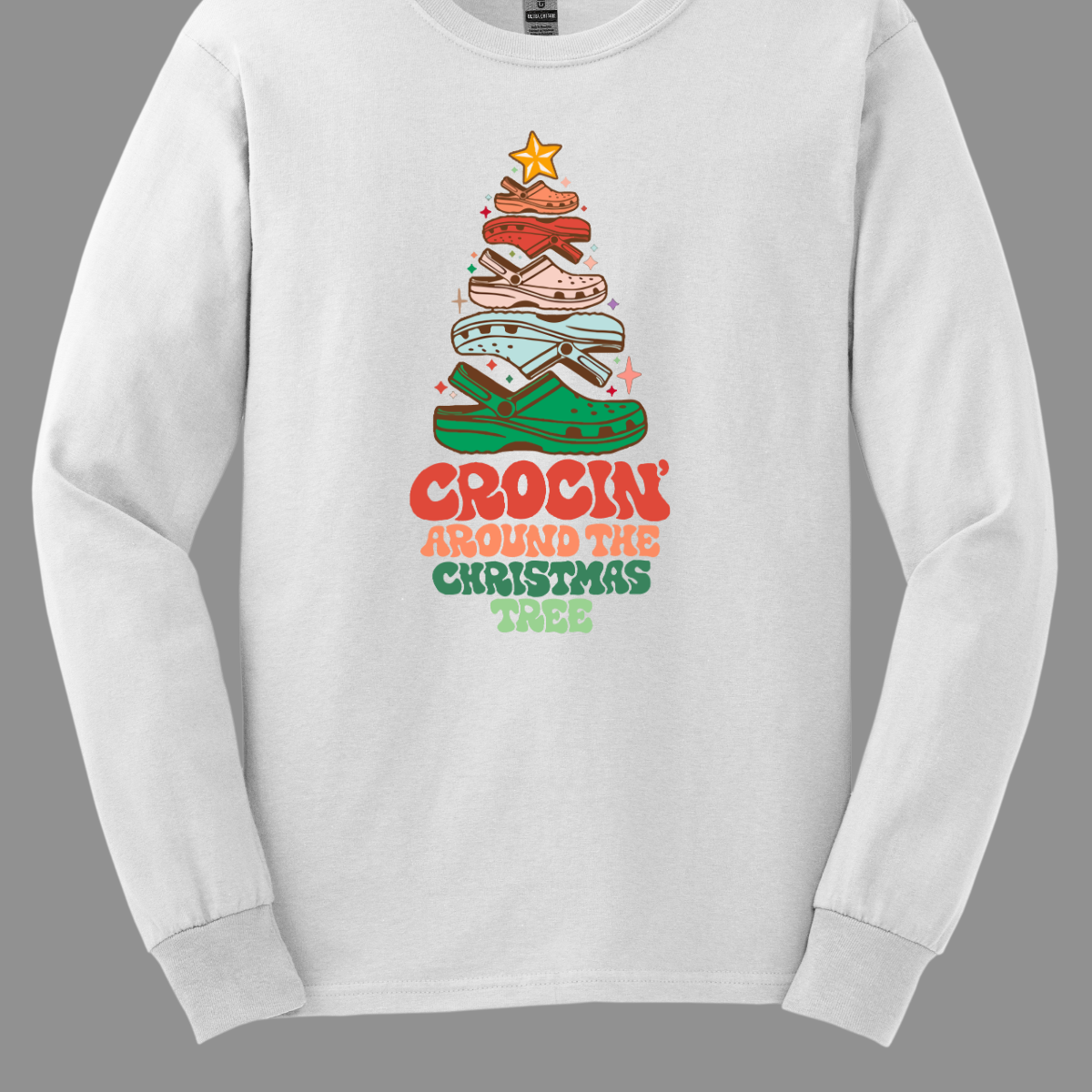 This fun t-shirt features the playful phrase "Crockin' Around the Christmas Tree" in a retro-inspired font that combines vibrant reds and greens for a festive touch. The centerpiece of the design is a whimsical Christmas tree crafted entirely from colorful croc shoes, stacked creatively to form the classic shape. Topping off the tree is a shiny star, adding a delightful finishing touch. Shop at Gorgeousware.com