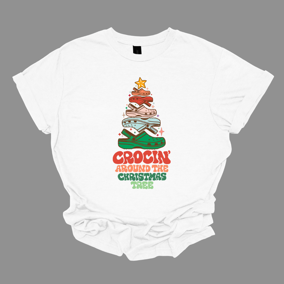 This fun t-shirt features the playful phrase "Crockin' Around the Christmas Tree" in a retro-inspired font that combines vibrant reds and greens for a festive touch. The centerpiece of the design is a whimsical Christmas tree crafted entirely from colorful croc shoes, stacked creatively to form the classic shape. Topping off the tree is a shiny star, adding a delightful finishing touch. Shop at Gorgeousware.com