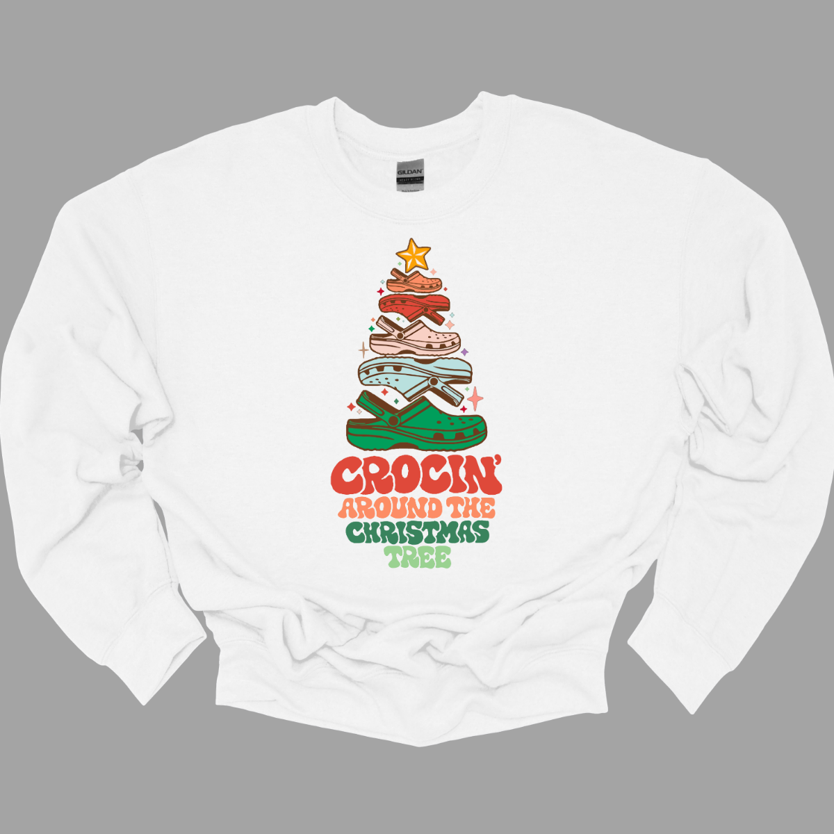 This fun t-shirt features the playful phrase "Crockin' Around the Christmas Tree" in a retro-inspired font that combines vibrant reds and greens for a festive touch. The centerpiece of the design is a whimsical Christmas tree crafted entirely from colorful croc shoes, stacked creatively to form the classic shape. Topping off the tree is a shiny star, adding a delightful finishing touch. Shop at Gorgeousware.com