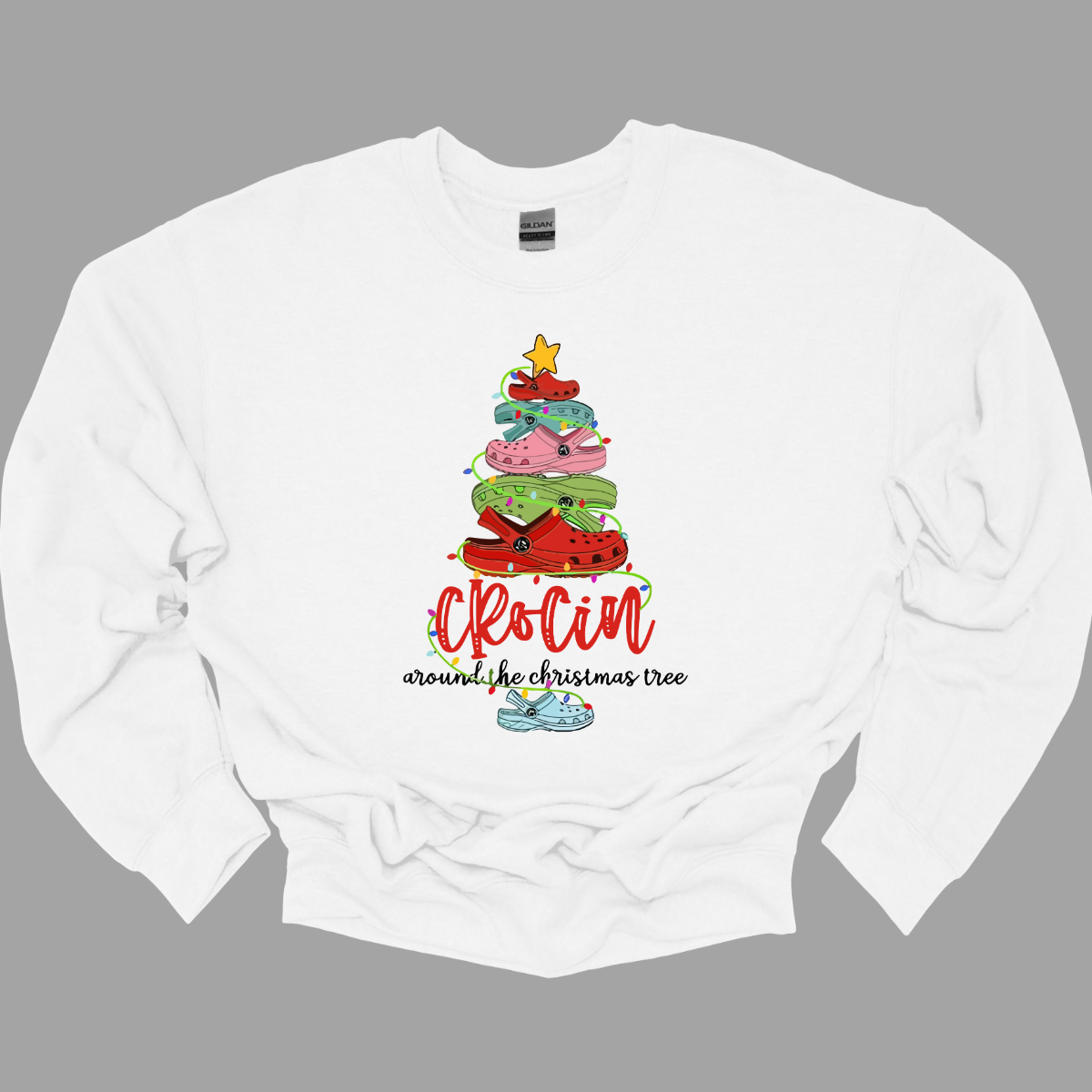 This playful t-shirt features the fun phrase "Crockin' Around the Christmas Tree" with a unique twist. The word "Crockin'" is showcased in a vibrant red cursive font, with playful spaces between the letters, adding a whimsical flair. Below that, the rest of the phrase is written in a smaller, elegant black cursive font, providing a charming contrast. Shop Gorgeousware.com