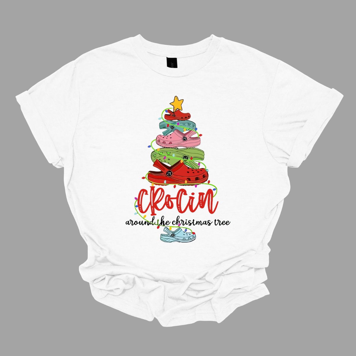 This playful t-shirt features the fun phrase "Crockin' Around the Christmas Tree" with a unique twist. The word "Crockin'" is showcased in a vibrant red cursive font, with playful spaces between the letters, adding a whimsical flair. Below that, the rest of the phrase is written in a smaller, elegant black cursive font, providing a charming contrast. Shop Gorgeousware.com