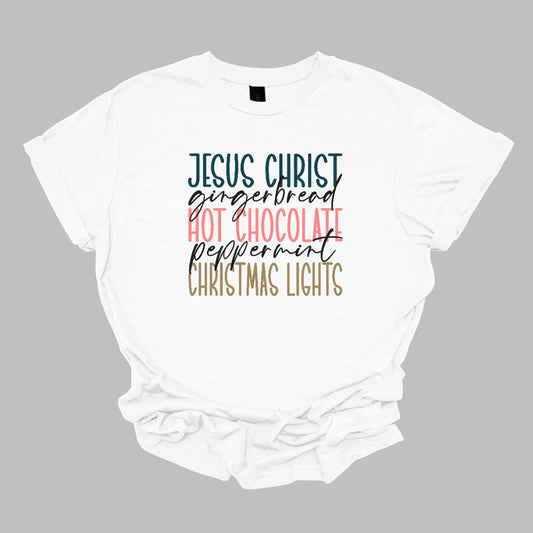 This festive t-shirt showcases a vibrant design with the phrase "JESUS CHRIST," "gingerbread," "HOT CHOCOLATE," "peppermint," and "CHRISTMAS LIGHTS" arranged vertically down the shirt. The words "JESUS CHRIST," "HOT CHOCOLATE," and "CHRISTMAS LIGHTS" are all in bold capital letters, each presented in a different bright color, creating a cheerful and eye-catching effect. Shop Gorgeousware.com