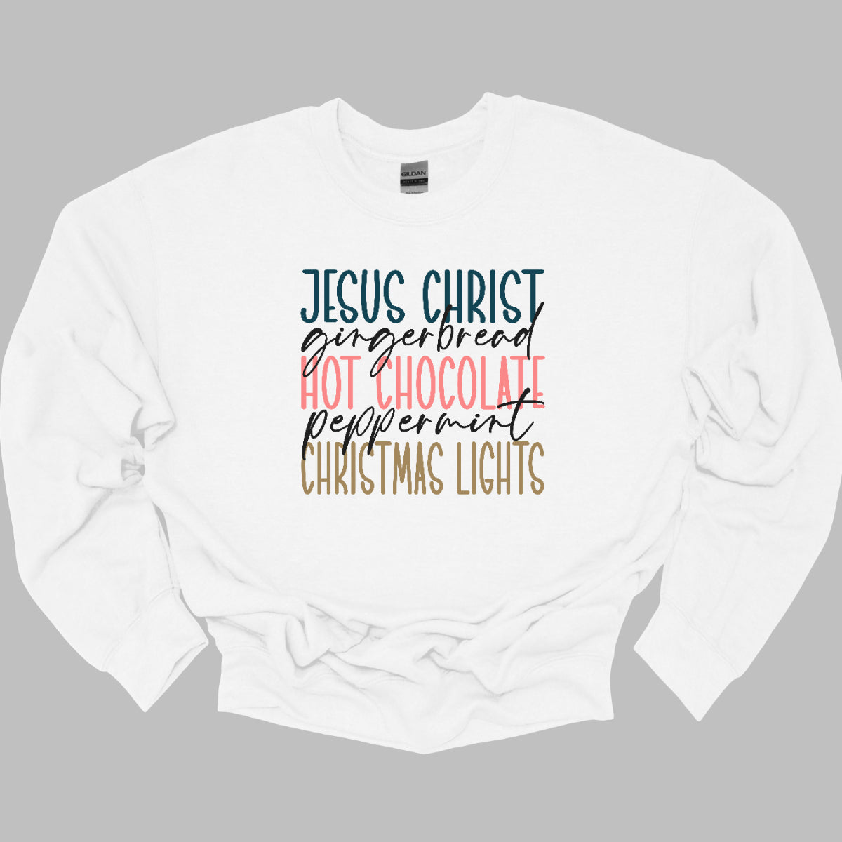 This festive t-shirt showcases a vibrant design with the phrase "JESUS CHRIST," "gingerbread," "HOT CHOCOLATE," "peppermint," and "CHRISTMAS LIGHTS" arranged vertically down the shirt. The words "JESUS CHRIST," "HOT CHOCOLATE," and "CHRISTMAS LIGHTS" are all in bold capital letters, each presented in a different bright color, creating a cheerful and eye-catching effect. Shop Gorgeousware.com