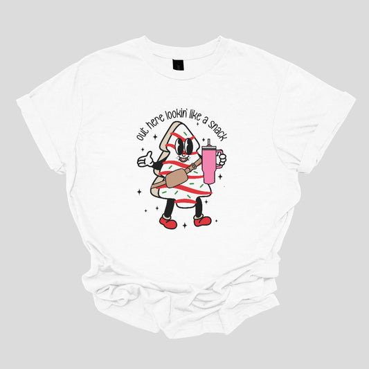 This playful t-shirt features the phrase "Out here lookin' like a snack" in a bold black stick figure font, giving it a fun and casual vibe. Beneath the text, a charming cartoon Christmas tree cake adds a whimsical touch, complete with a cheerful smiling face, a stylish belt bag, and a cup. The surrounding sparkles enhance the festive spirit, making this shirt a delightful choice for spreading holiday cheer with a humorous twist! Shop Gorgeousware.com