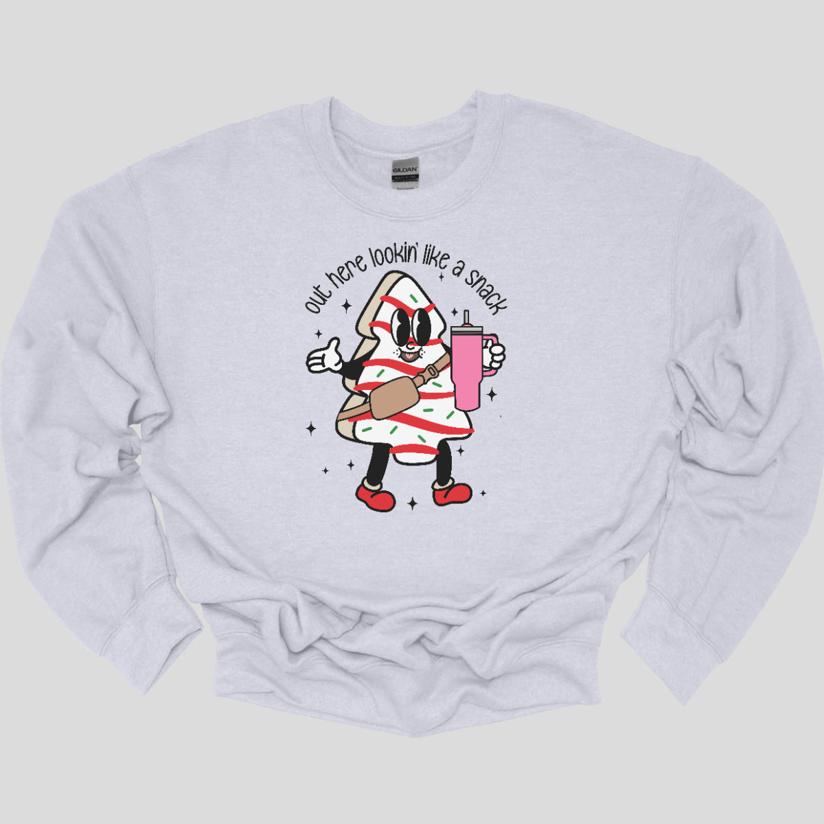 This playful t-shirt features the phrase "Out here lookin' like a snack" in a bold black stick figure font, giving it a fun and casual vibe. Beneath the text, a charming cartoon Christmas tree cake adds a whimsical touch, complete with a cheerful smiling face, a stylish belt bag, and a cup. The surrounding sparkles enhance the festive spirit, making this shirt a delightful choice for spreading holiday cheer with a humorous twist! Shop Gorgeousware.com