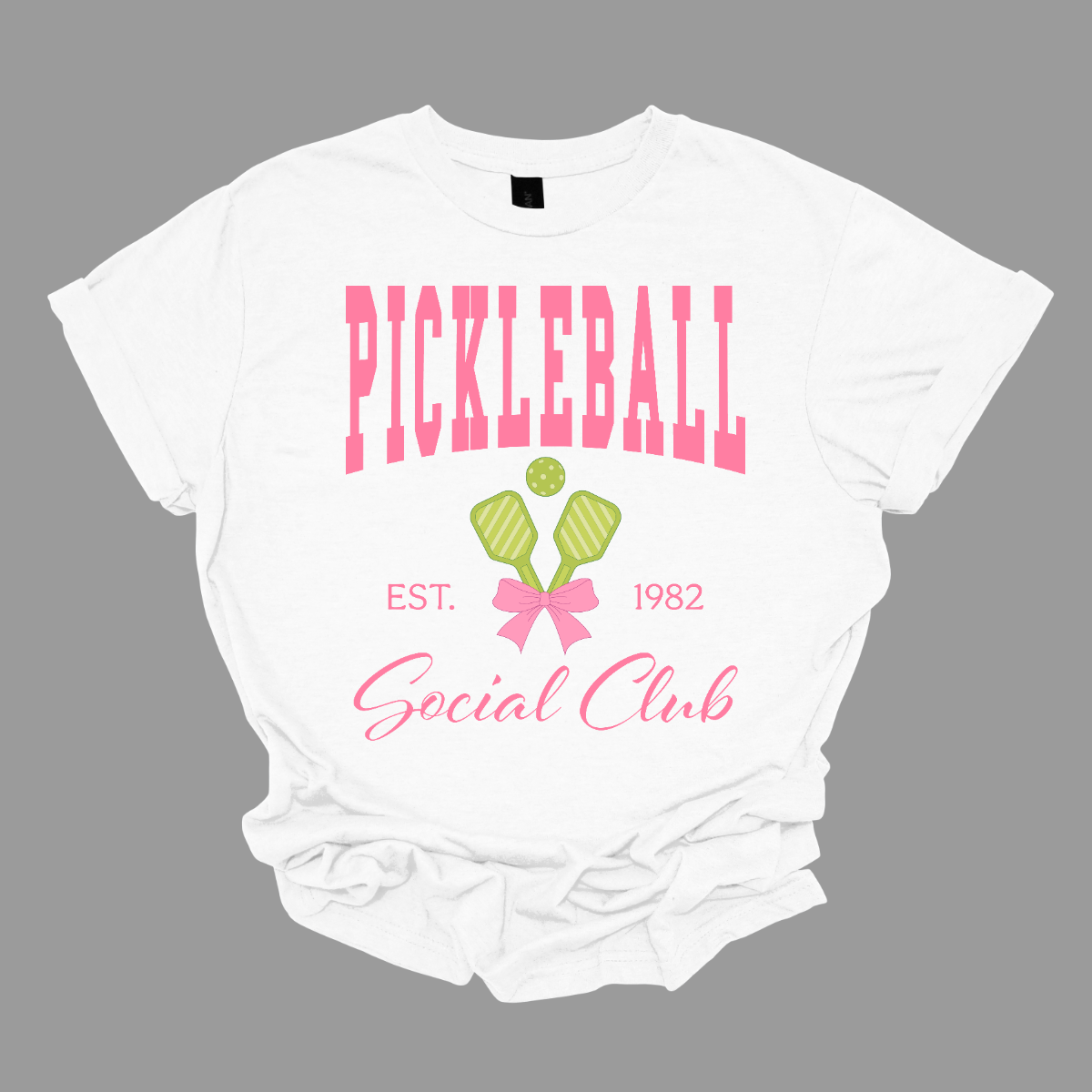 The shirt design features the phrase "Pickleball Social Club" in bold, hot pink font, giving it a fun and vibrant look. Below, "Est. 1982" is displayed in a stylish, slightly smaller font, adding a touch of vintage charm.
Two pickleball paddles are crossed in the center, creating a dynamic focal point, with a pickleball placed between them to emphasize the sport. A pink bow is tied around the intersection of the paddles, adding a playful and fashionable touch. Shop Gorgeousware.com
