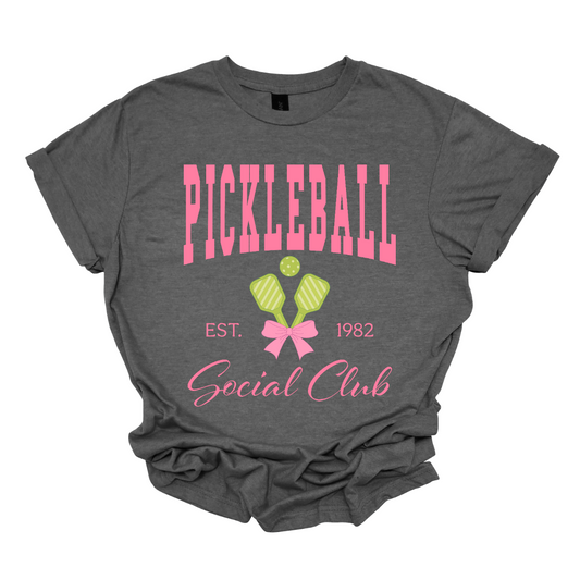The shirt design features the phrase "Pickleball Social Club" in bold, hot pink font, giving it a fun and vibrant look. Below, "Est. 1982" is displayed in a stylish, slightly smaller font, adding a touch of vintage charm.
Two pickleball paddles are crossed in the center, creating a dynamic focal point, with a pickleball placed between them to emphasize the sport. A pink bow is tied around the intersection of the paddles, adding a playful and fashionable touch.  Shop Gorgeousware.com