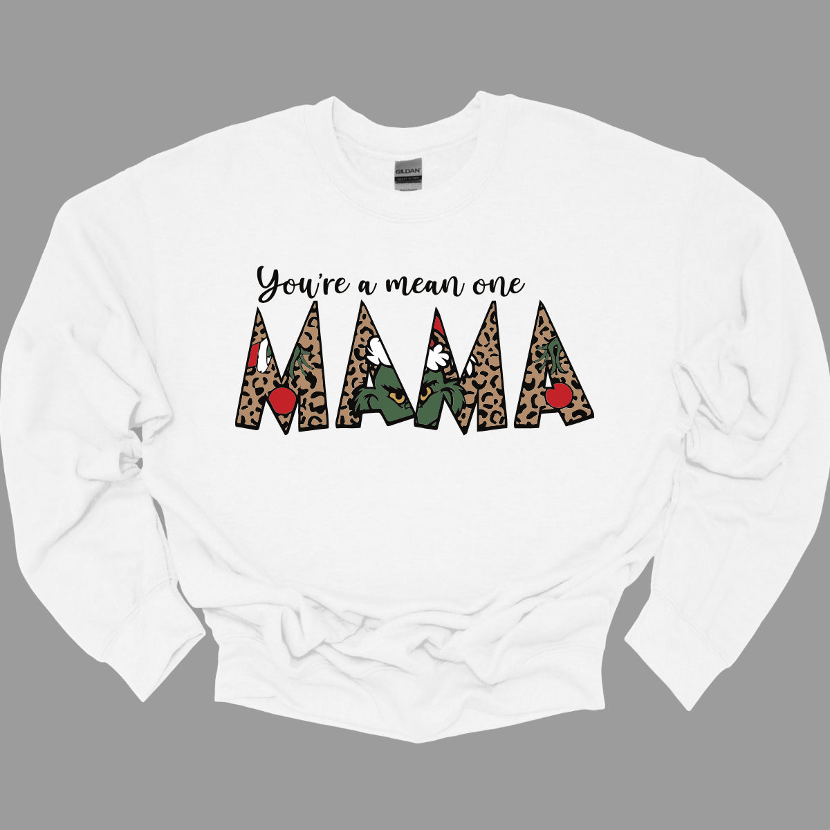 The phrase "You're a Mean One Mama" takes center stage, with "You're a Mean One" written in elegant black cursive font that flows beautifully across the fabric. The standout element is the word "Mama," which is rendered in bold, eye-catching letters. Each letter of "Mama" is filled with a quirky depiction of the Grinch, set against a vibrant cheetah print background. Shop Gorgeousware.com