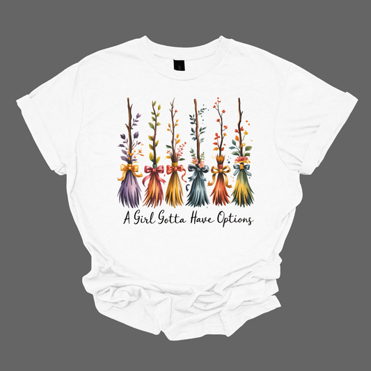 Add a little magic to your fall wardrobe with this fun and whimsical Halloween-inspired tee! The design features six wispy brooms lined up in playful autumn colors, each with its own unique charm. Beneath them, the phrase “A Girl Gotta Have Options” is written, making this shirt a cheeky and stylish nod to witchy fall vibes. Shop at Gorgeousware.com