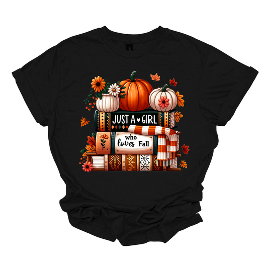 Bring cozy fall vibes to your wardrobe with this autumn-inspired graphic tee! The design features two pumpkins and a small pot of flowers perched on a stack of books with the spines reading “Just a Girl” and “Who Loves Fall.” An orange-and-white scarf wraps around the books while leaves gently fall, creating the perfect scene for autumn lovers. Shop at Gorgeousware.com