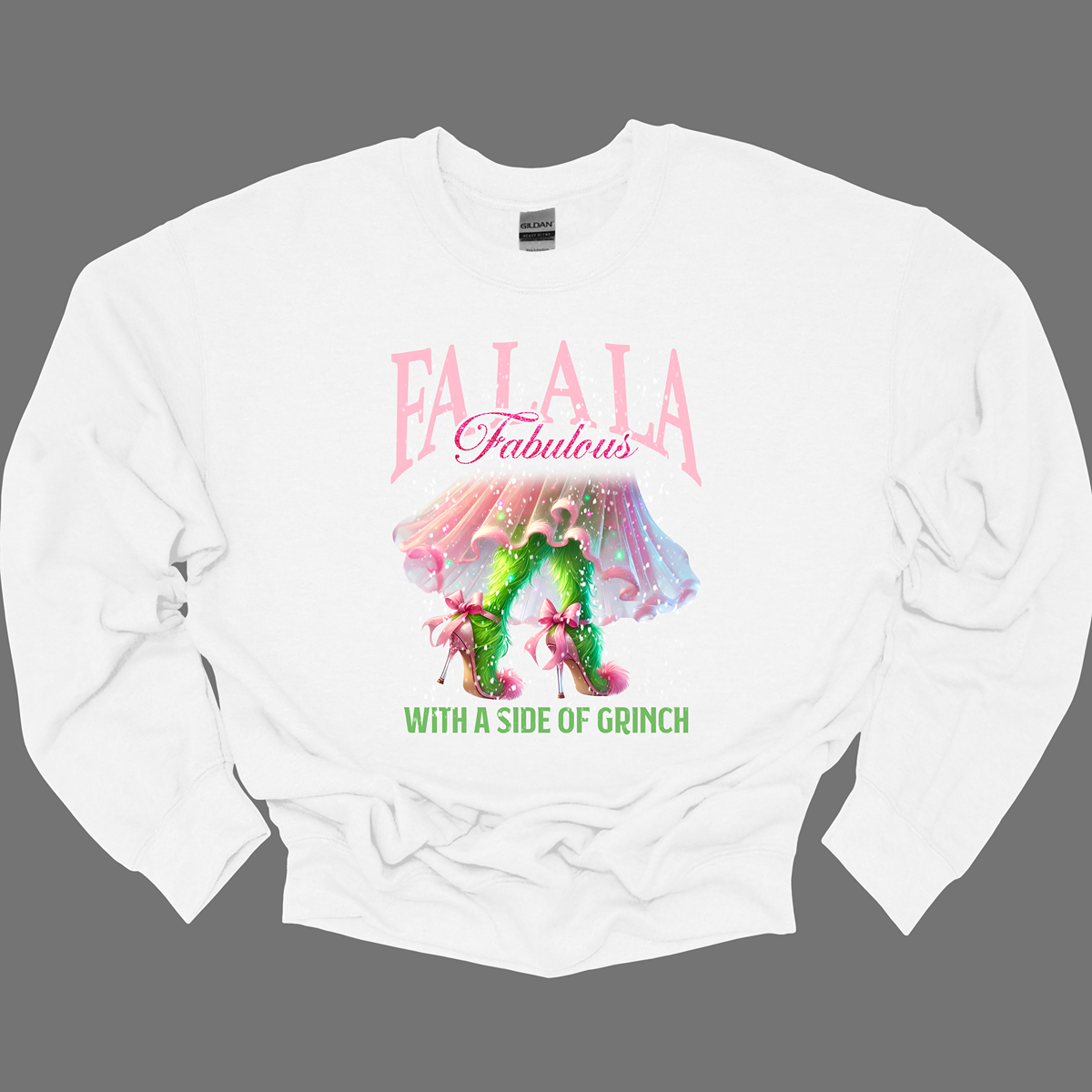 The design features a playful and festive take on the Grinch character with a stylish twist. The phrase "Fa La La Fabulous" is at the top in a fun, cursive font with a bouncy, holiday-inspired feel. The word "Fabulous" stands out in bold, glamorous lettering with subtle sparkles, giving it a chic vibe. Below, the Grinch is shown with her girly green legs, styled in light pink high heels adorned with oversized bows at the back, adding a sweet and fashionable touch. Shop at Gorgeousware.com