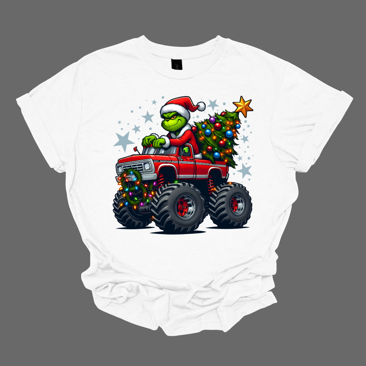 White t-shirt with a graphic of the grinch driving a truck that is hauling a Christmas tree, on agrey background. Shop at Gorgeousware.com