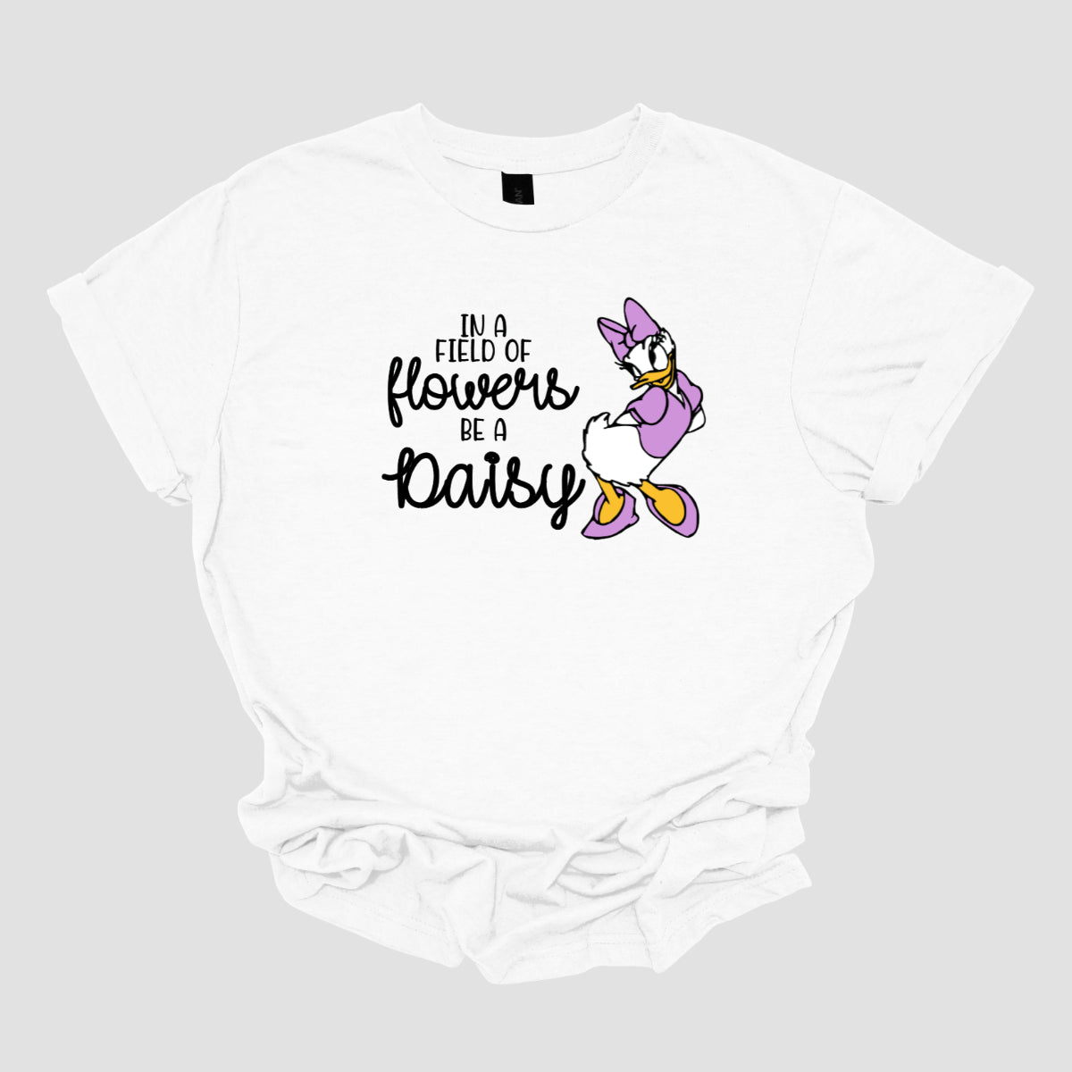 This t-shirt features an inspiring and charming design with the uplifting message: "In a Field of Flowers Be a Daisy." The phrase is styled with a delightful mix of fonts. The words "In a Field of" and "Be a" are written in a playful black font, bringing a touch of fun to the overall look. The words "Flowers" and "Daisy" are highlighted in a graceful black cursive font, adding a touch of elegance and flow. Next to the text, there’s a cute illustration of the cartoon character Daisy. Shop Gorgeousware.com