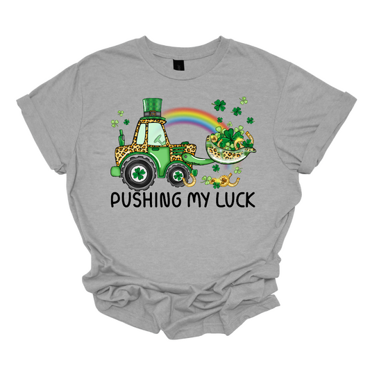 A playful and whimsical garment that captures the spirit of the holiday with a touch of fun. This shirt features a lively design with a tractor playfully shoveling a load of shamrocks, creating a unique and charming representation of pushing one's luck in a St. Patrick's Day context.  The design showcases a tractor adorned with shamrocks, symbolizing the act of "pushing" luck in a light-hearted and festive manner. Shop at Gorgeousware.com