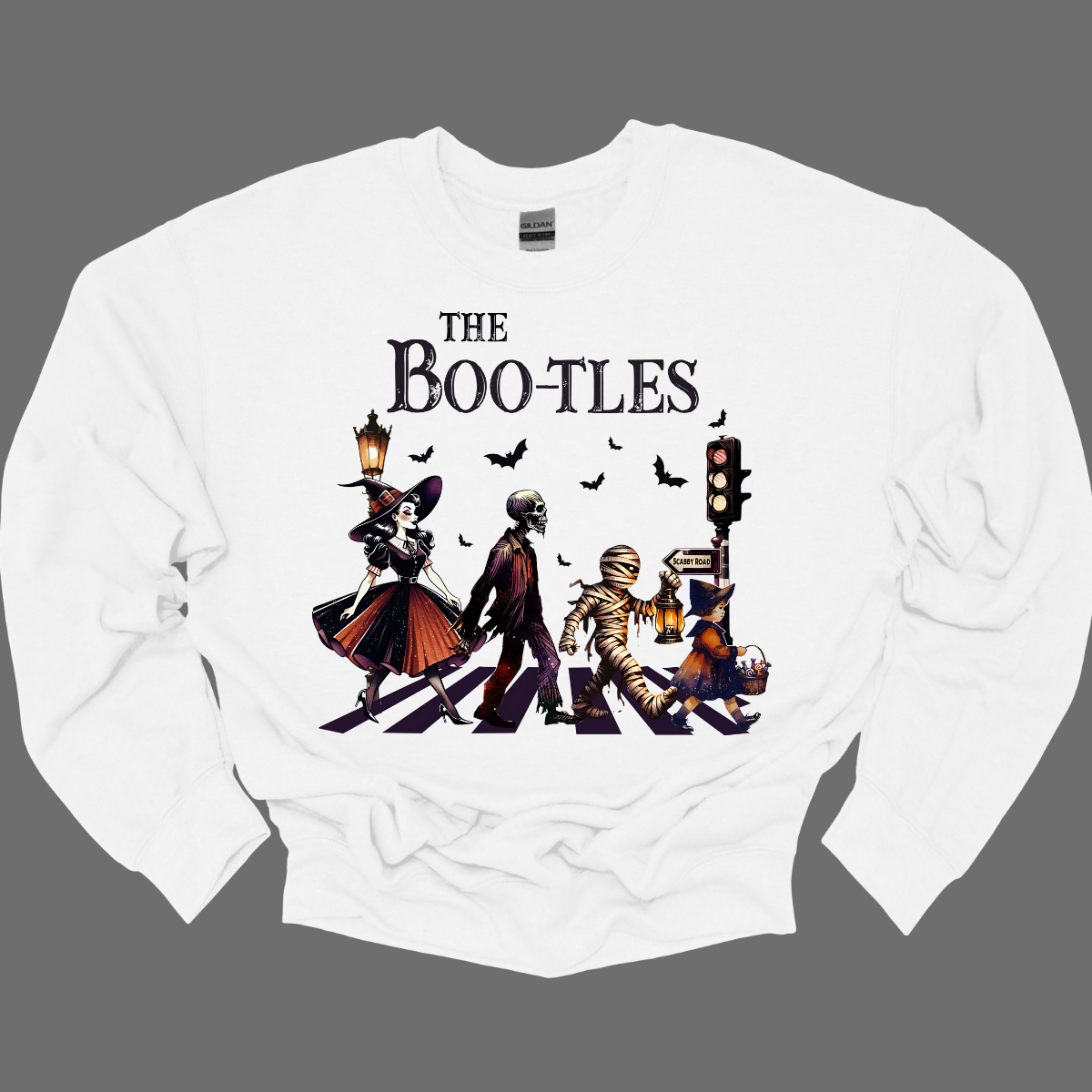 Step into a spooky twist on a classic with our 'Boot-les' tee! This Halloween-themed shirt takes inspiration from the iconic Abbey Road album cover and reimagines it with a creepy-cool lineup of Halloween characters. Picture this: a mischievous witch, a spooky skeleton, a creepy mummy, and a charming little witch, all crossing a spectral road in style.  Shop at Gorgeousware.com